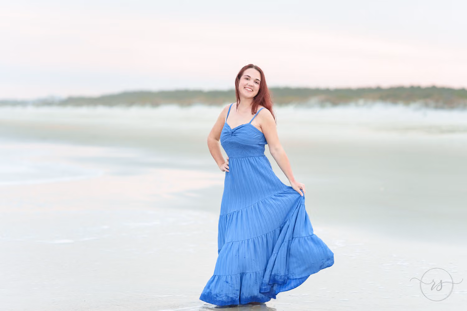 Senior swishing her long blue dress on the flat sand near the water at sunset at Huntington Beach State Park