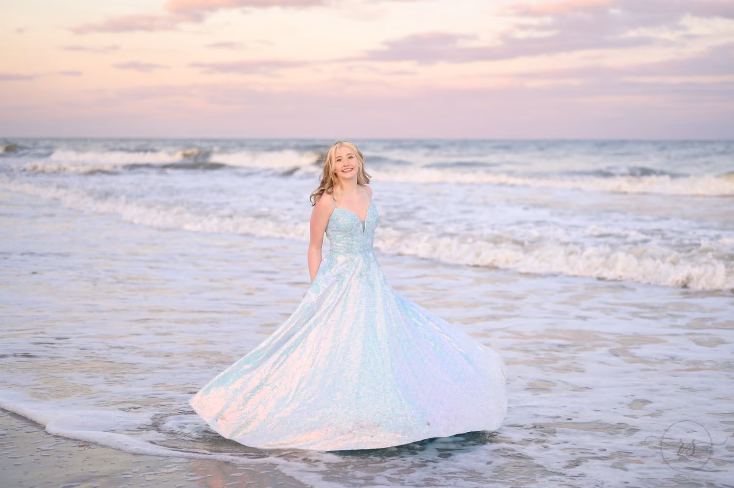 Teen twirling her shimmering blue prom gown at the edge of the surf under a pastel sky at Huntington Beach State Park