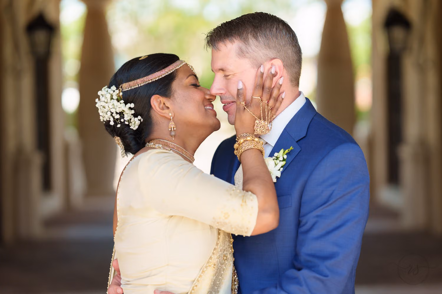 Bride in traditional dress and jewelry holding the groom's face as they touch noses under the columns at 21 Main Events