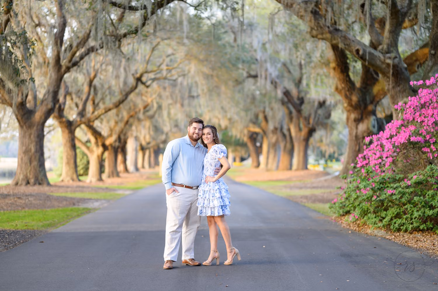 Couple standing together on a road under a long canopy of mossy oaks with pink azaleas at Caledonia Golf and Fish Club