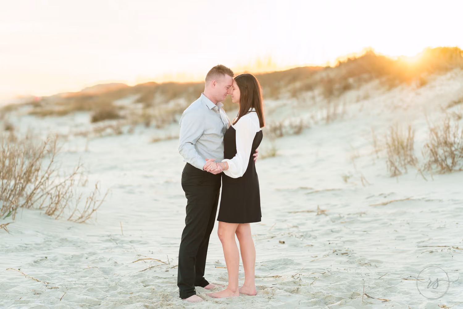 Couple standing close in the white sand dunes as the sun sets behind the sea oats at Huntington Beach State Park