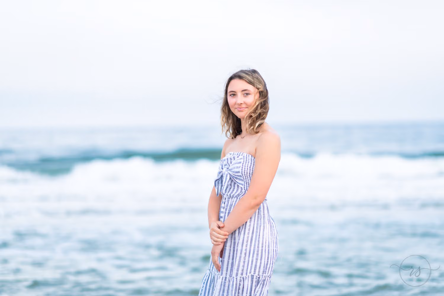 Senior in a striped dress standing on the beach with waves breaking behind her at Huntington Beach State Park