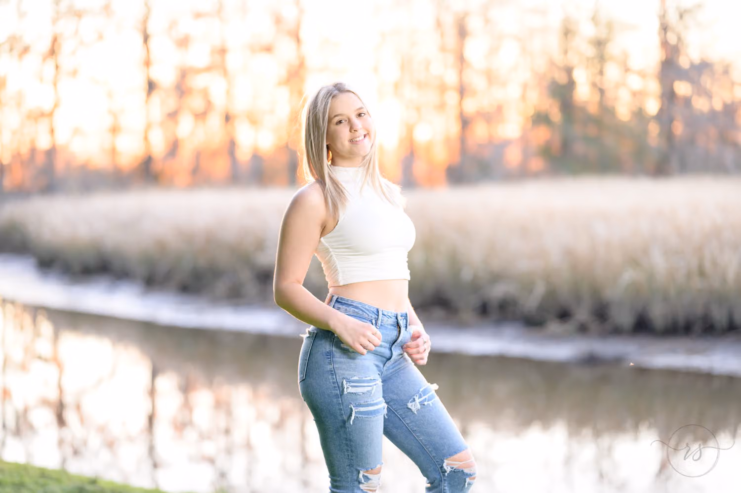 Senior girl in ripped jeans and a white top standing by the marsh creek in soft evening light at Caledonia Golf & Fish Club