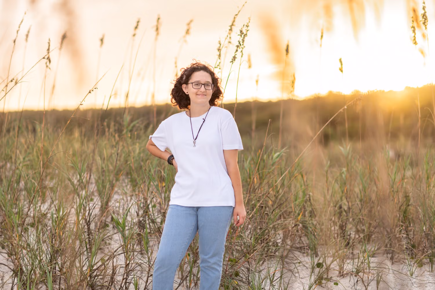 Senior in a white tee standing in tall sea oats with the sunset glowing behind her at Huntington Beach State Park