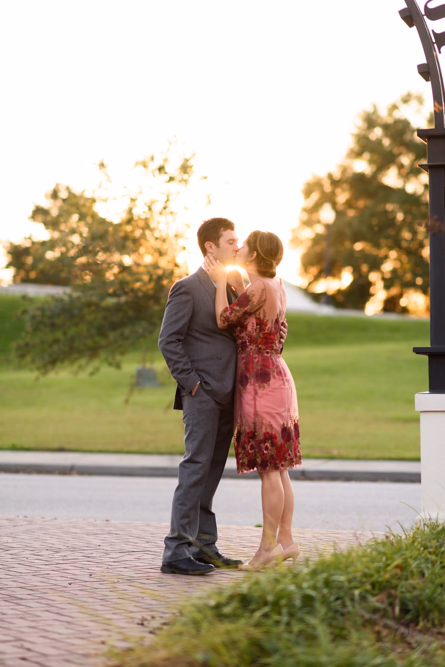 Couple kissing on a brick sidewalk with warm sunlight glowing through the trees behind them along the Conway Riverwalk