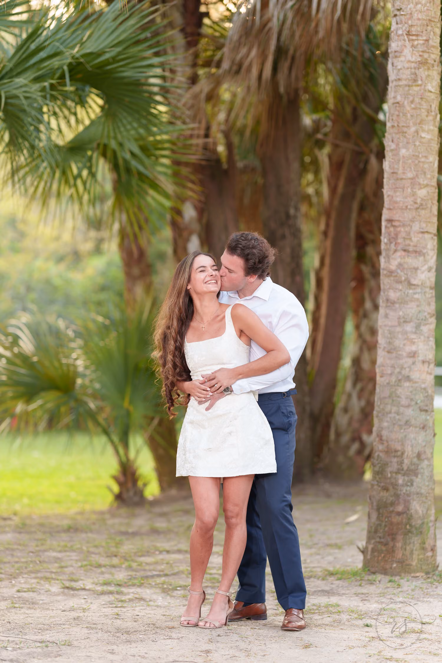 Man hugging his fiancée from behind and kissing her cheek among palm trees in late sunlight at Huntington Beach State Park