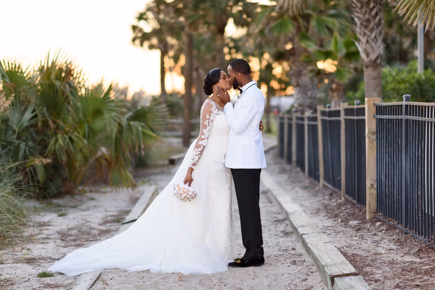 Groom in a white jacket kissing the bride on a sandy path lined with palms at the DoubleTree Resort in Myrtle Beach
