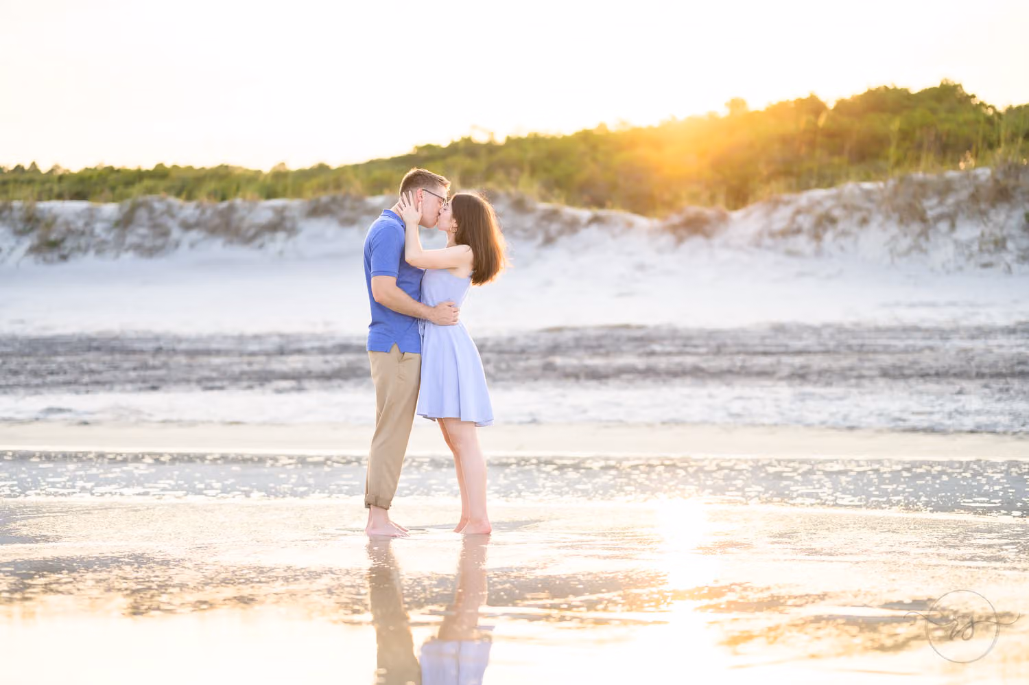 Newly engaged couple kissing at the water's edge with their reflection and the dunes at sunset at Huntington Beach State Park