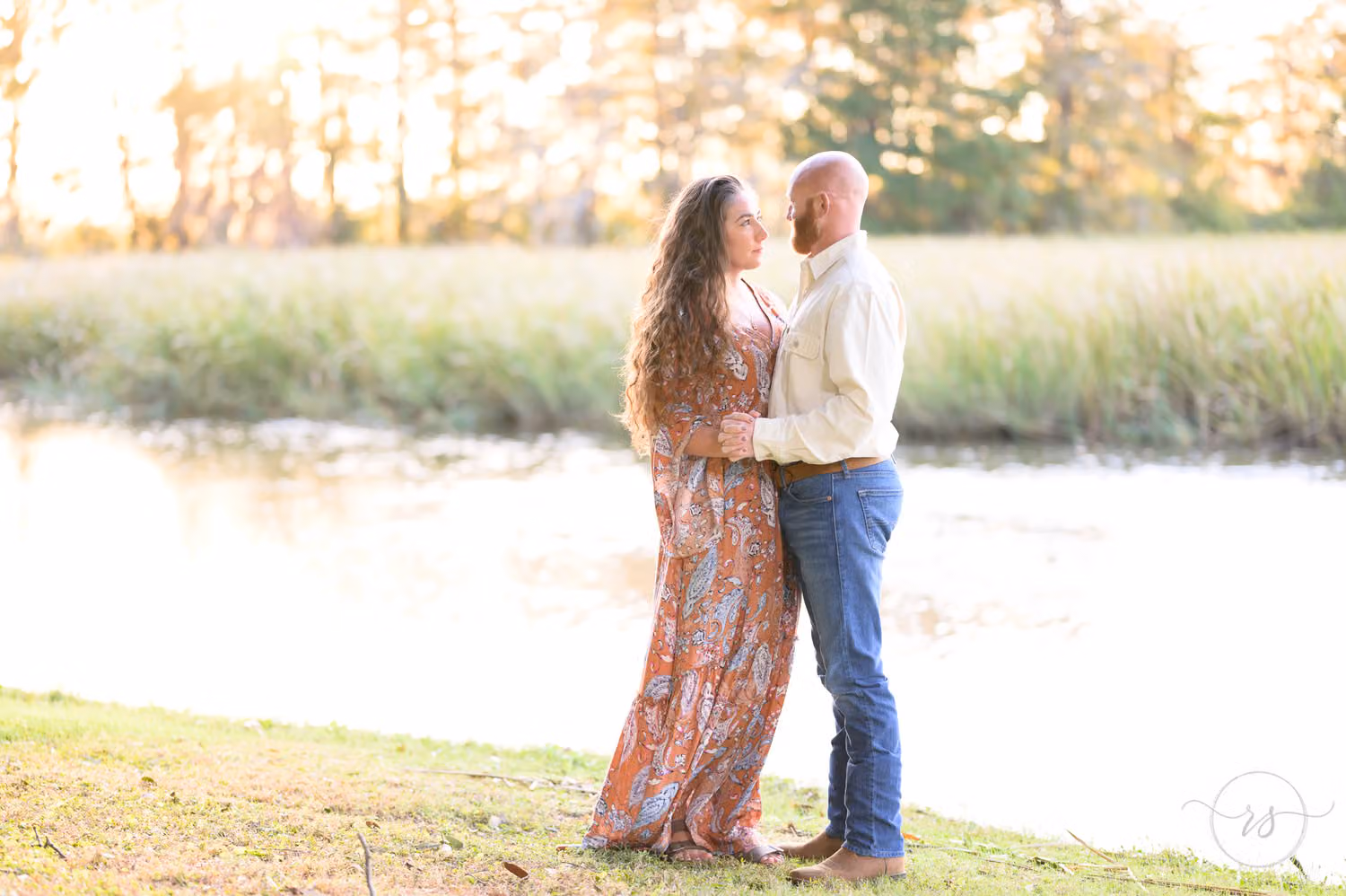 Couple standing close on the riverbank with marsh grass glowing in the sunset at Caledonia Golf & Fish Club