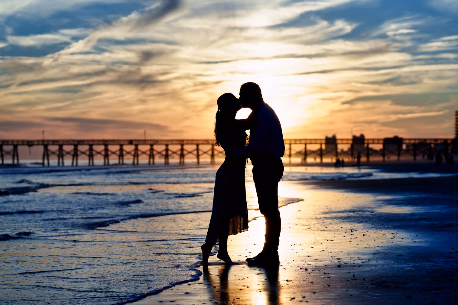 Silhouette of a couple about to kiss on the beach with the pier and a colorful sunset behind them at Cherry Grove Point