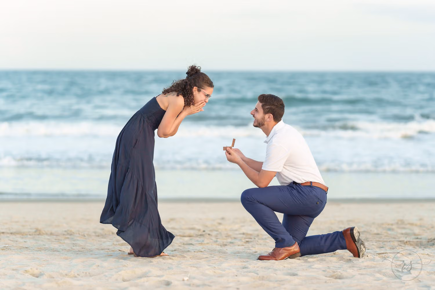 Man on one knee as his girlfriend in a navy dress doubles over in surprise on the beach at Huntington Beach State Park