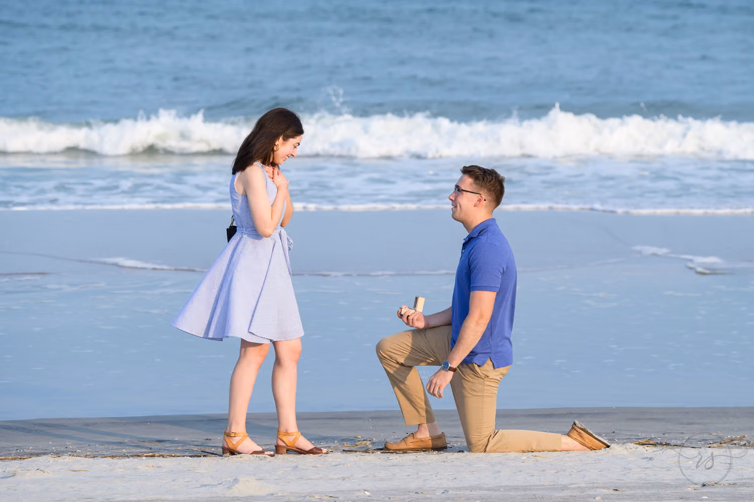 Man on one knee with a ring as his girlfriend in lavender covers her mouth at Huntington Beach State Park