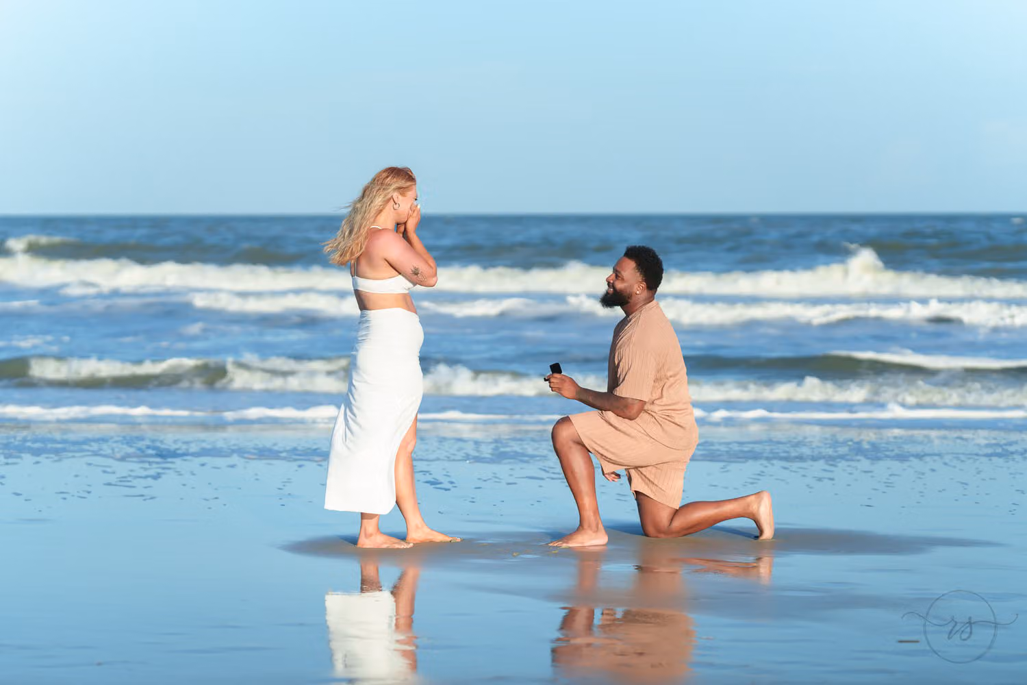 Man kneeling in the shallow surf with a ring box as his girlfriend in white reacts at Huntington Beach State Park