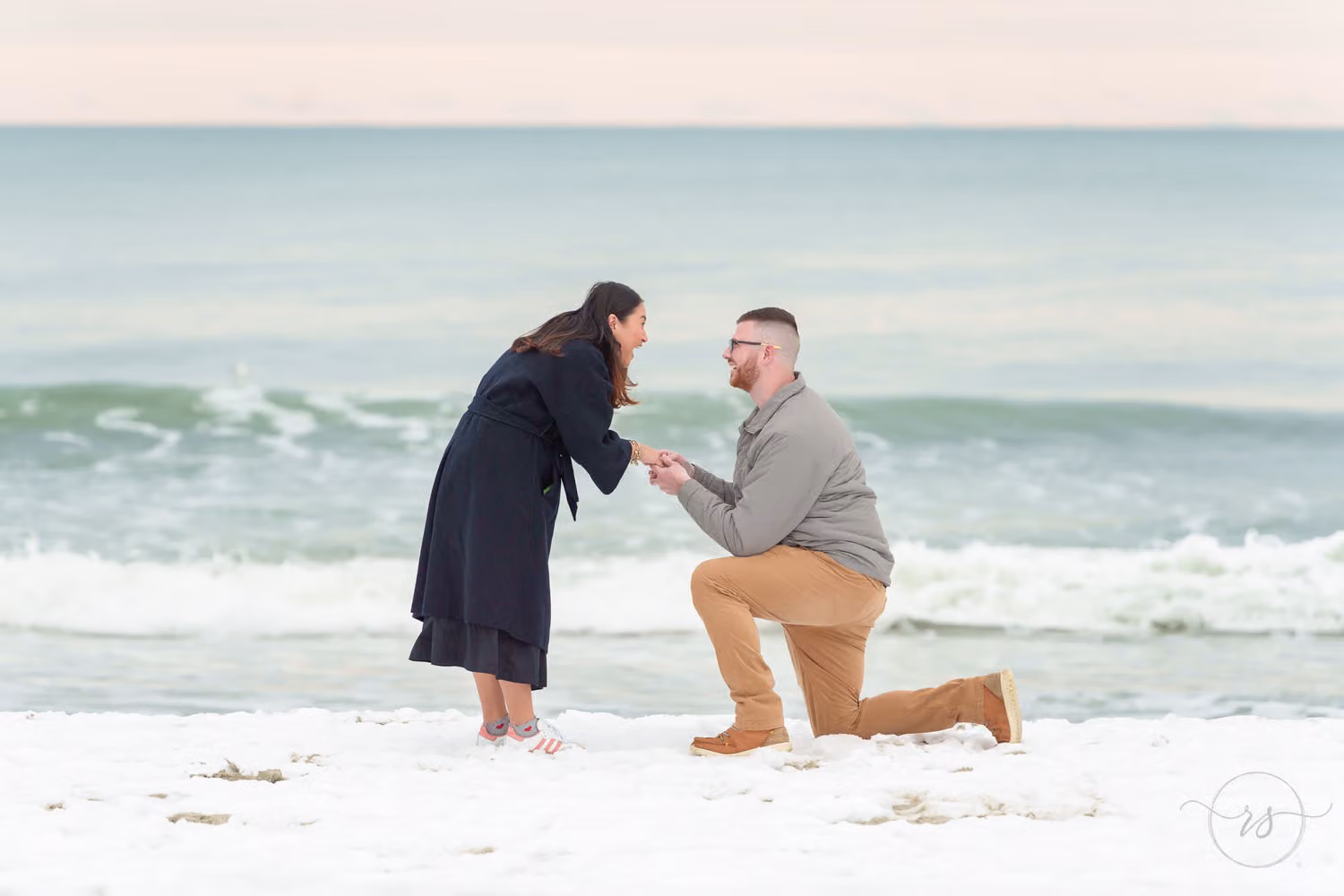 Man on one knee holding her hands on a snow-covered beach as she leans in smiling on Garden City Beach