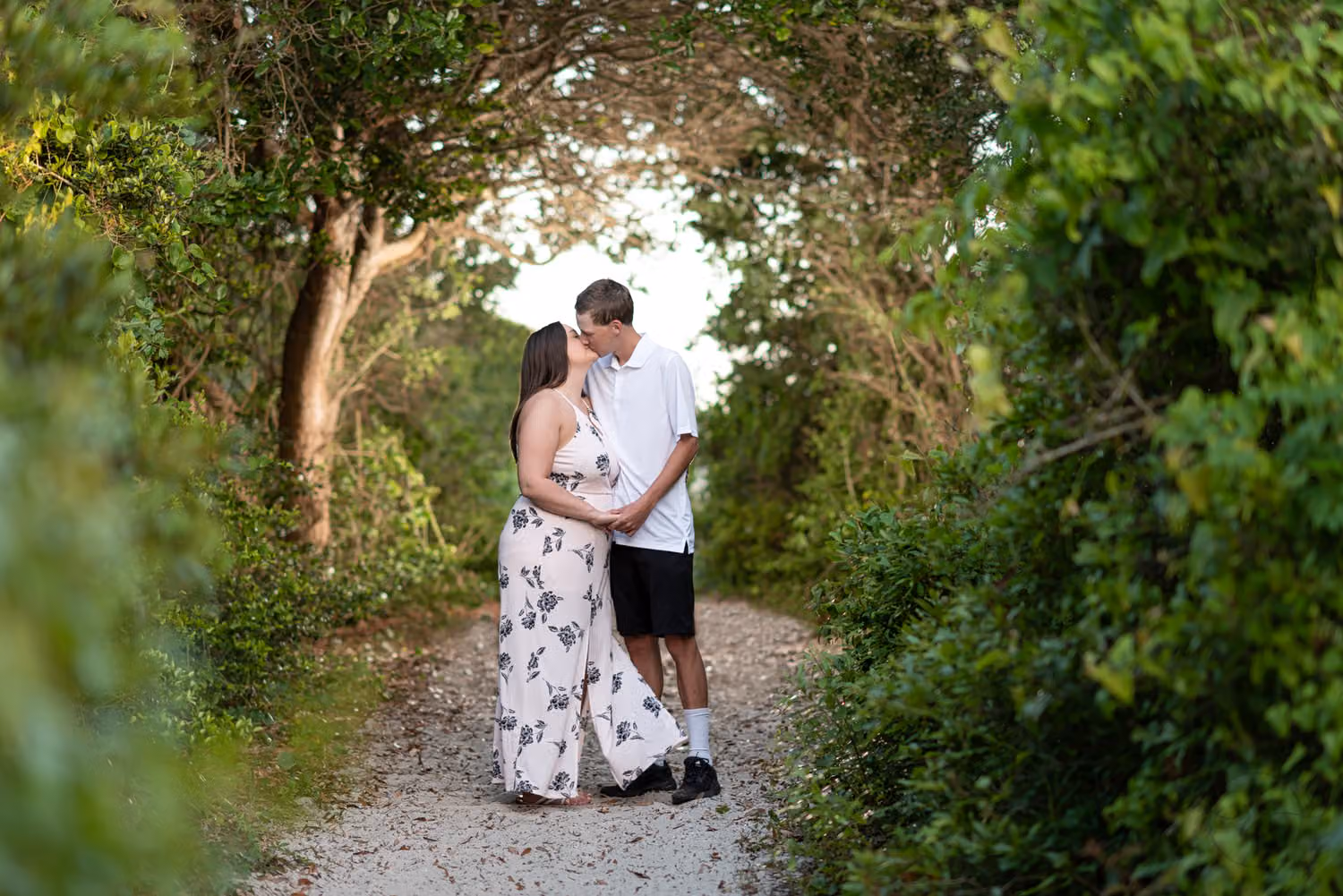 Couple kissing on a sandy path under a tunnel of dark trees lit from behind at Huntington Beach State Park