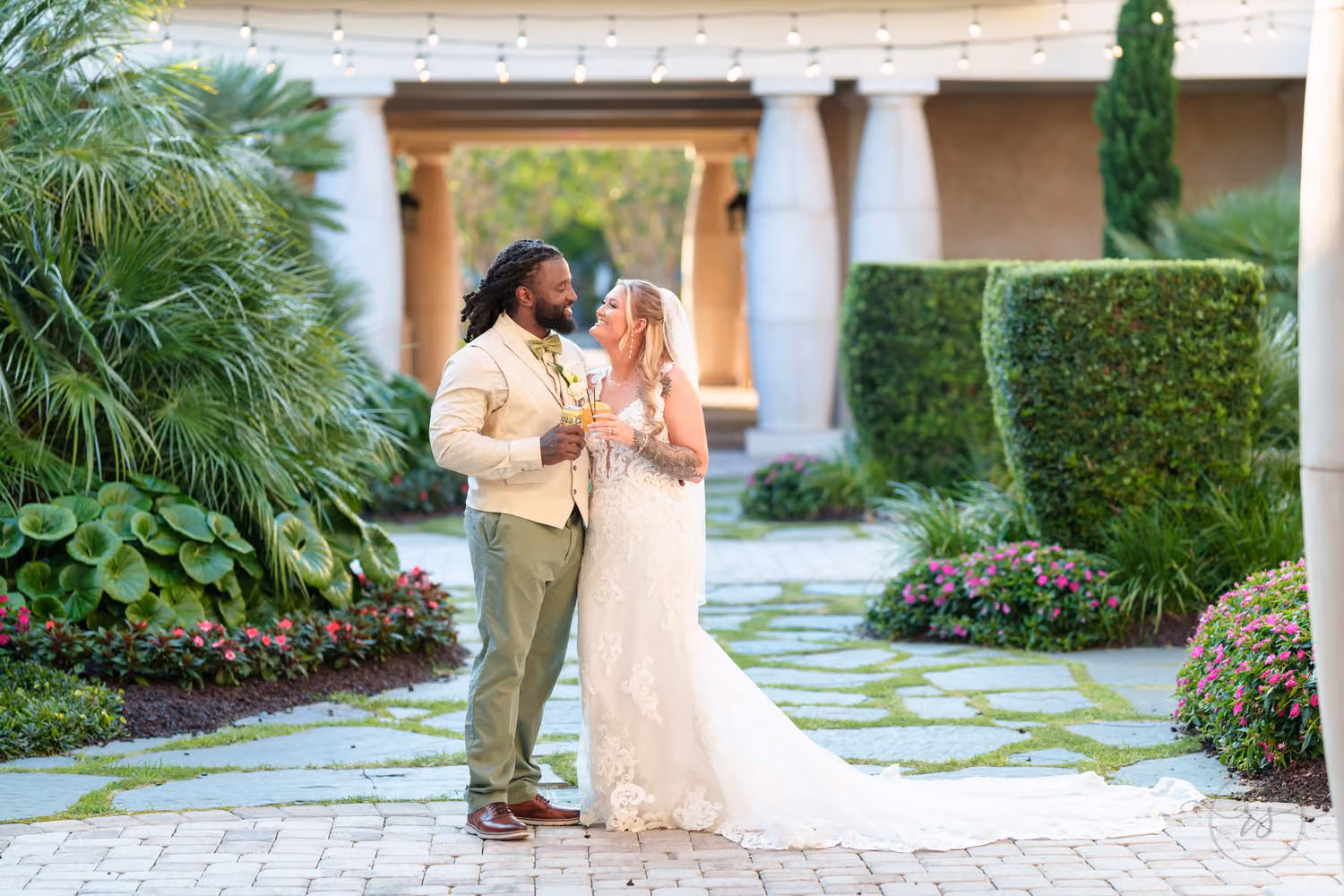 Groom in a tan jacket and bride raising champagne glasses in the columned courtyard garden at 21 Main Events