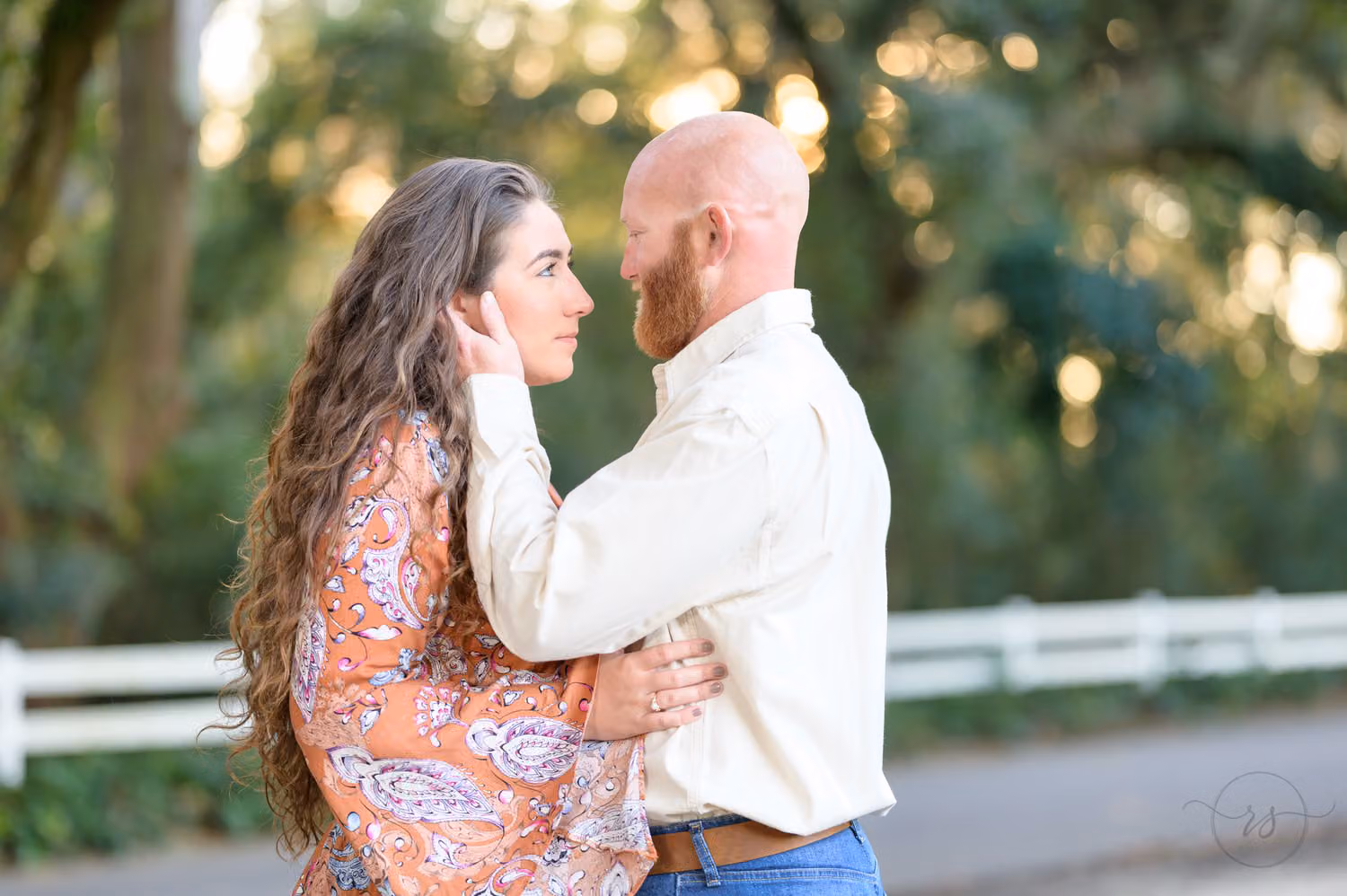Man touching his fiancée's face as she looks up at him beside a white fence and oak trees at Caledonia Golf & Fish Club