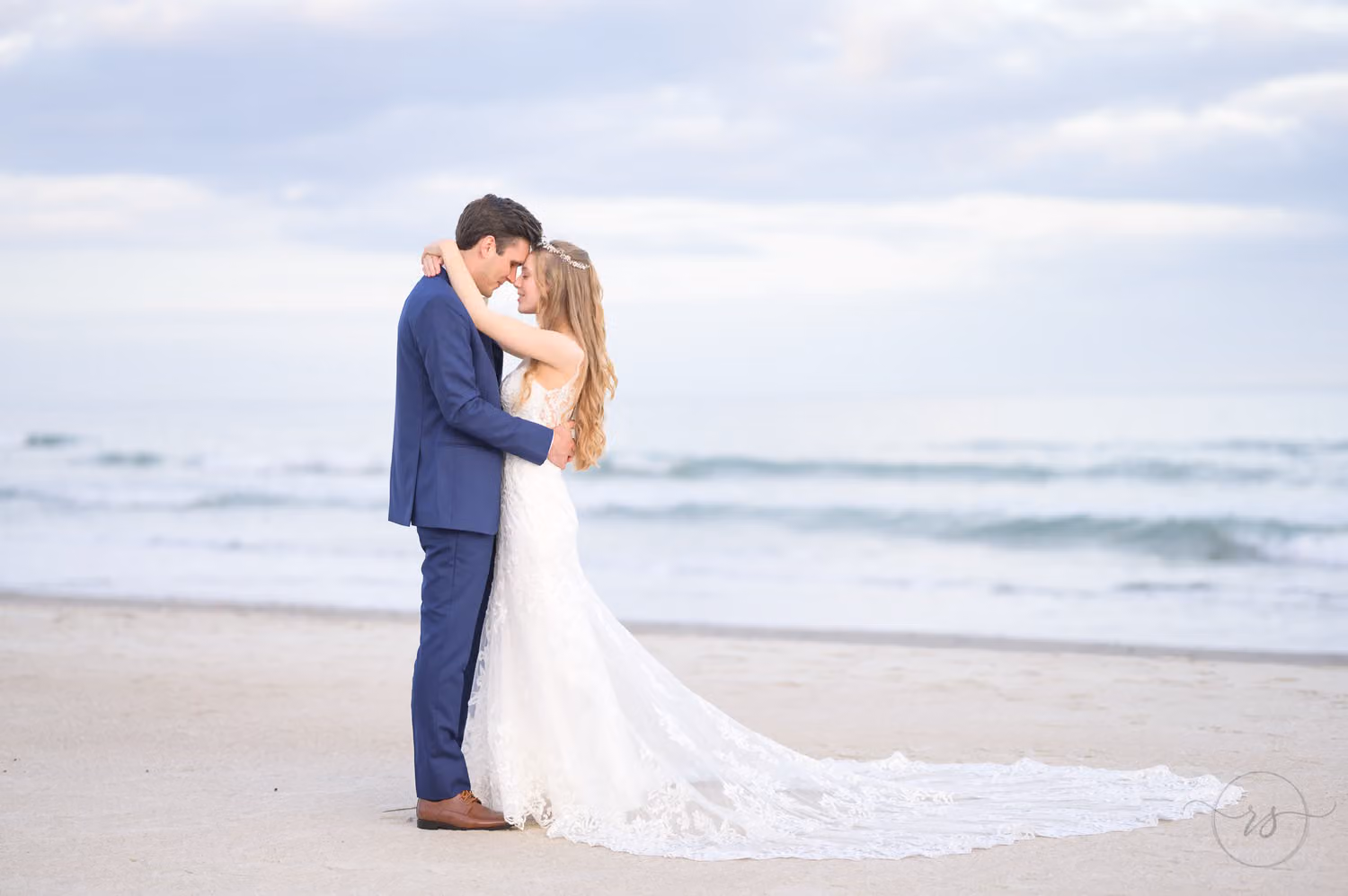 Bride and groom in a blue suit touching foreheads on the beach with waves rolling in behind them in Pawleys Island