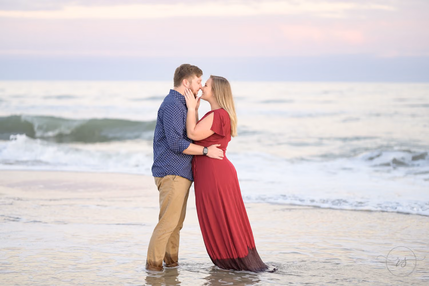 Couple touching noses in shallow water at sunset, her red gown trailing in the surf at Huntington Beach State Park