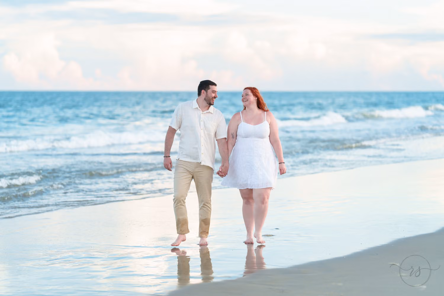 Engaged couple walking hand in hand on the wet sand and smiling at each other as waves roll in