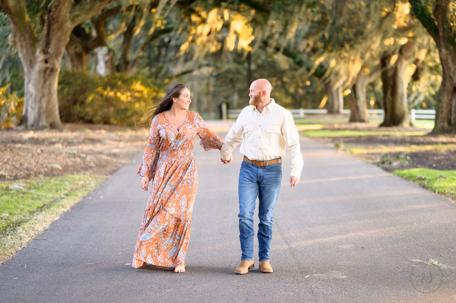 Couple holding hands and walking down a road under oaks draped in Spanish moss at Caledonia Golf & Fish Club