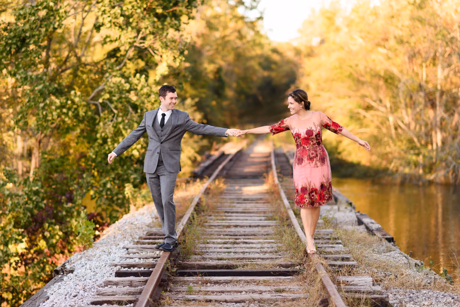 Couple holding hands while balancing on the rails of an old train track through fall trees along the Conway Riverwalk