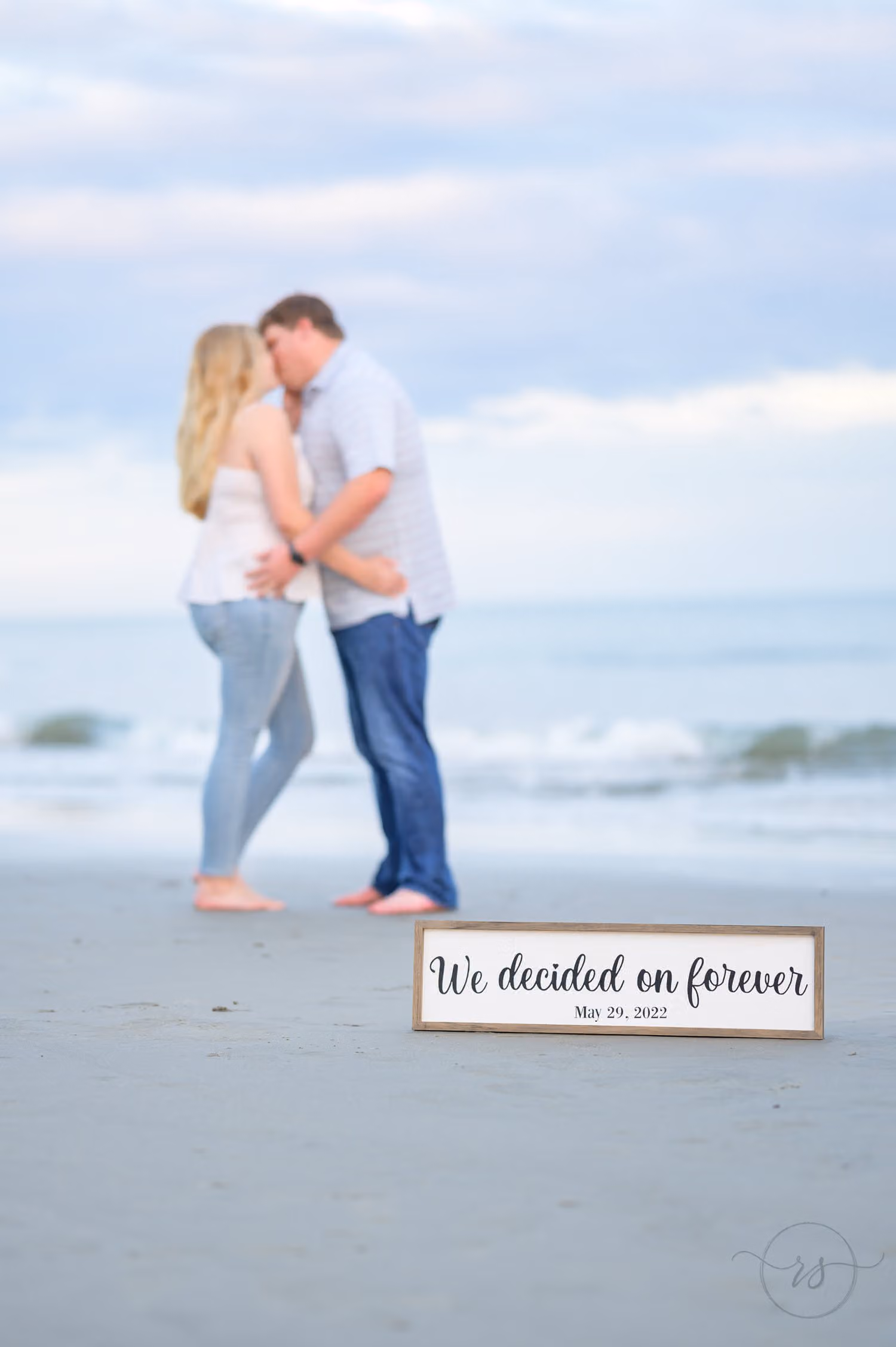 Couple kissing at the water's edge behind a we decided on forever sign in the sand at Huntington Beach State Park