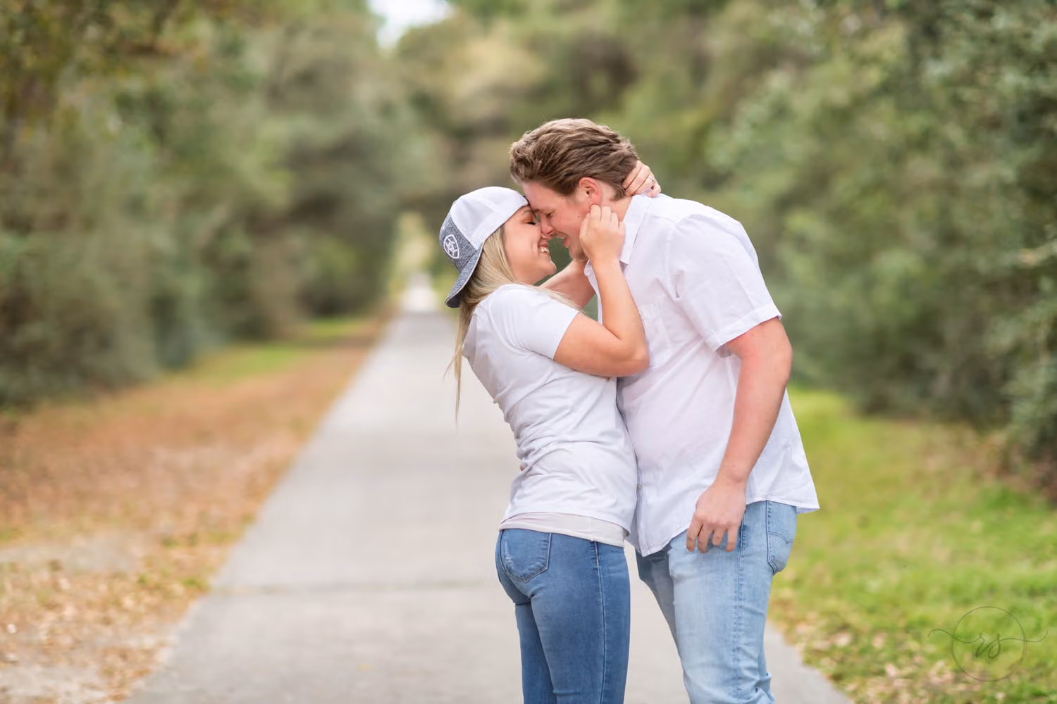 Woman wearing her fiancé's baseball cap backward as they kiss on a tree-lined path at Huntington Beach State Park