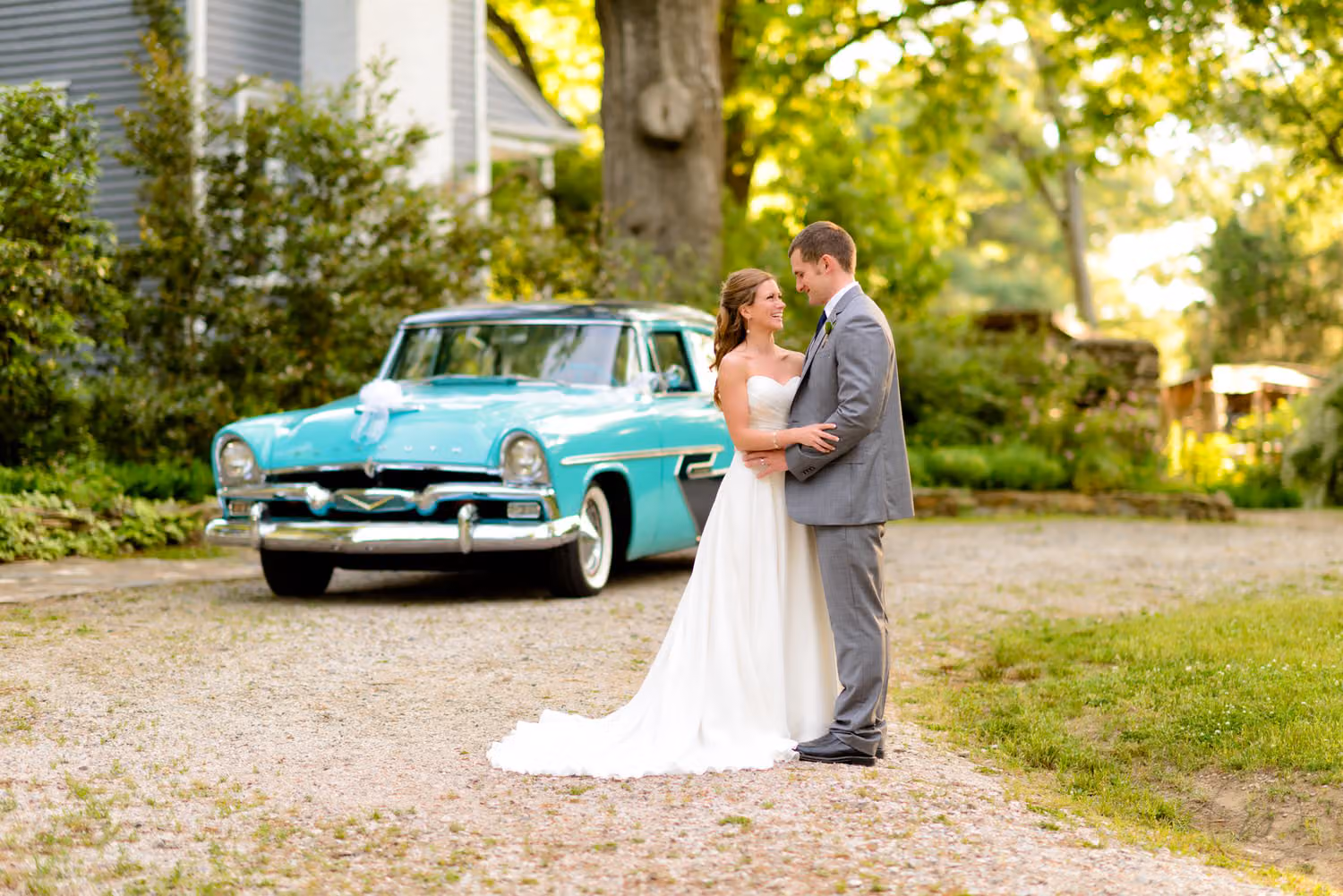Bride and groom in a gray suit hugging beside a turquoise vintage car on a shady gravel drive at The Ivy Place