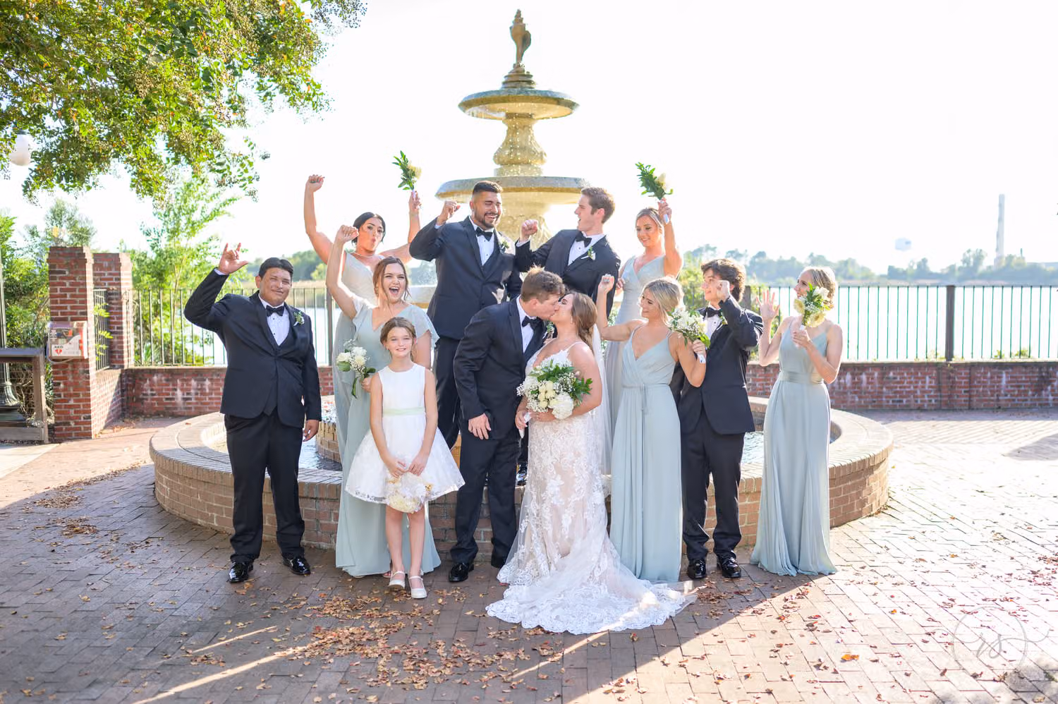 Bridesmaids in pale blue and groomsmen cheering as the bride and groom kiss by a garden fountain at the Kaminski House Museum