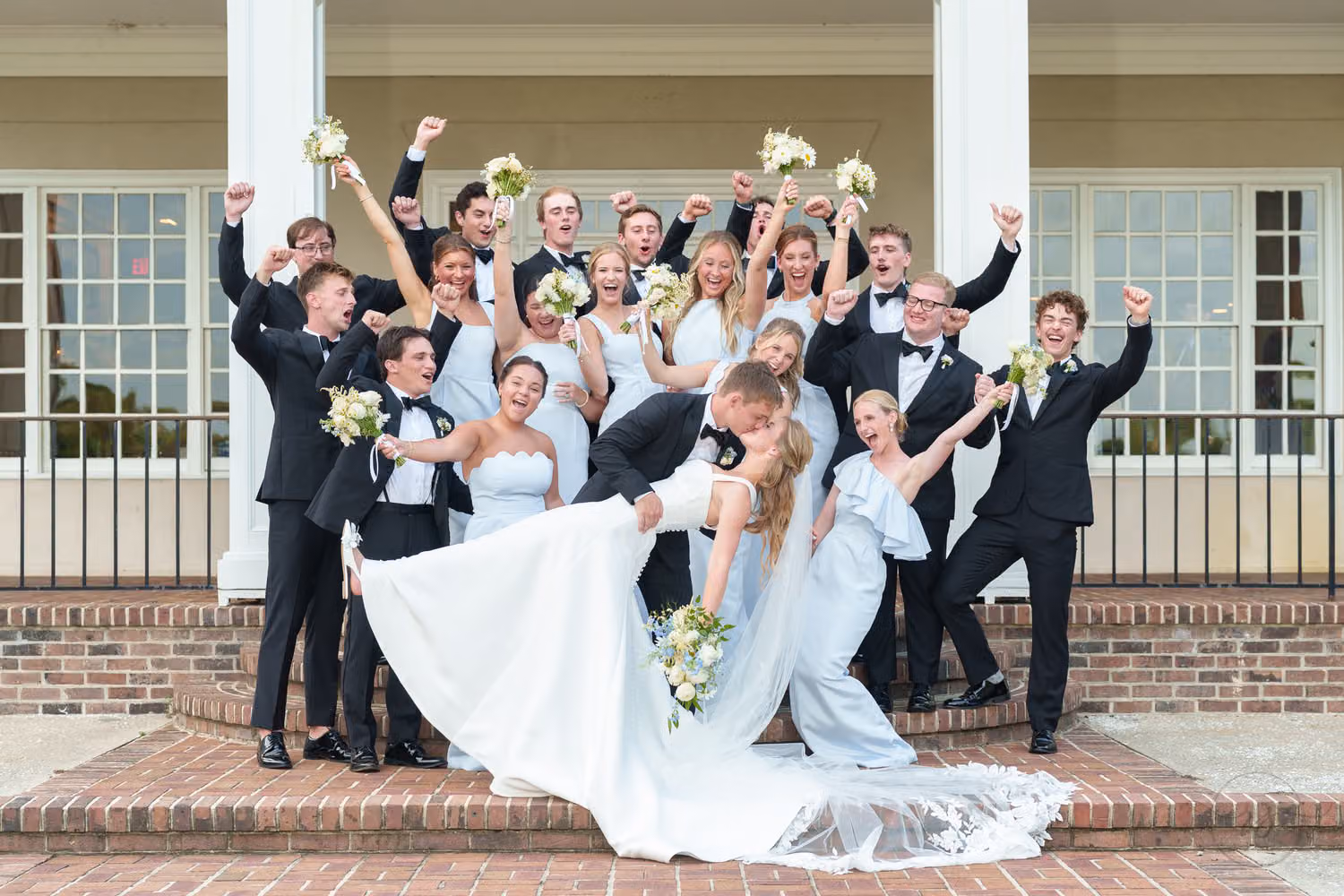 Groom dipping the bride on the clubhouse steps as the large wedding party cheers with bouquets raised at Pawleys Plantation