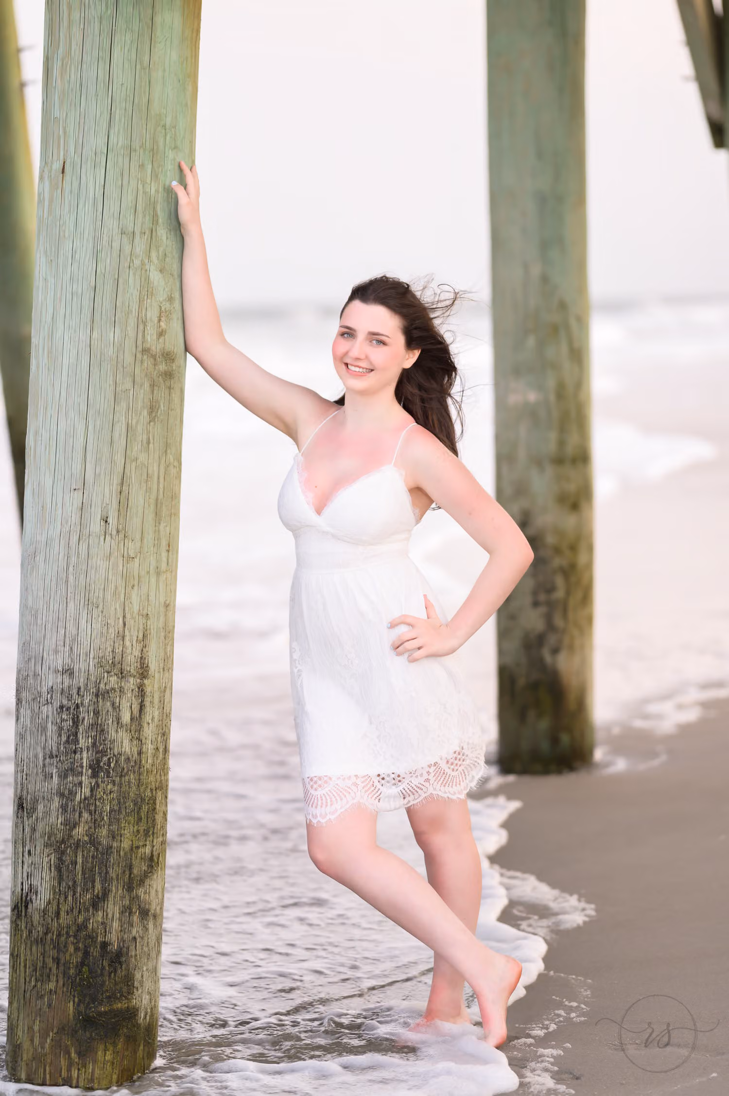 Senior in a white dress with her hair blowing, standing between pier pilings in the surf at Myrtle Beach State Park
