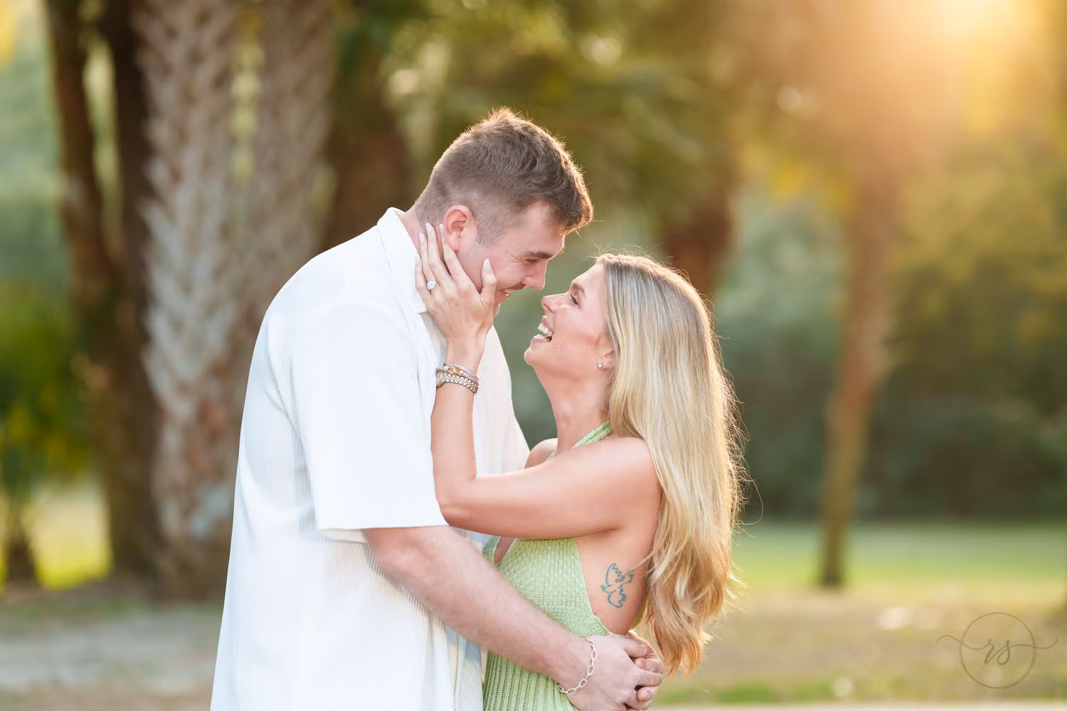 Man holding his fiancée's face as she smiles up at him under palm trees in golden light at Huntington Beach State Park