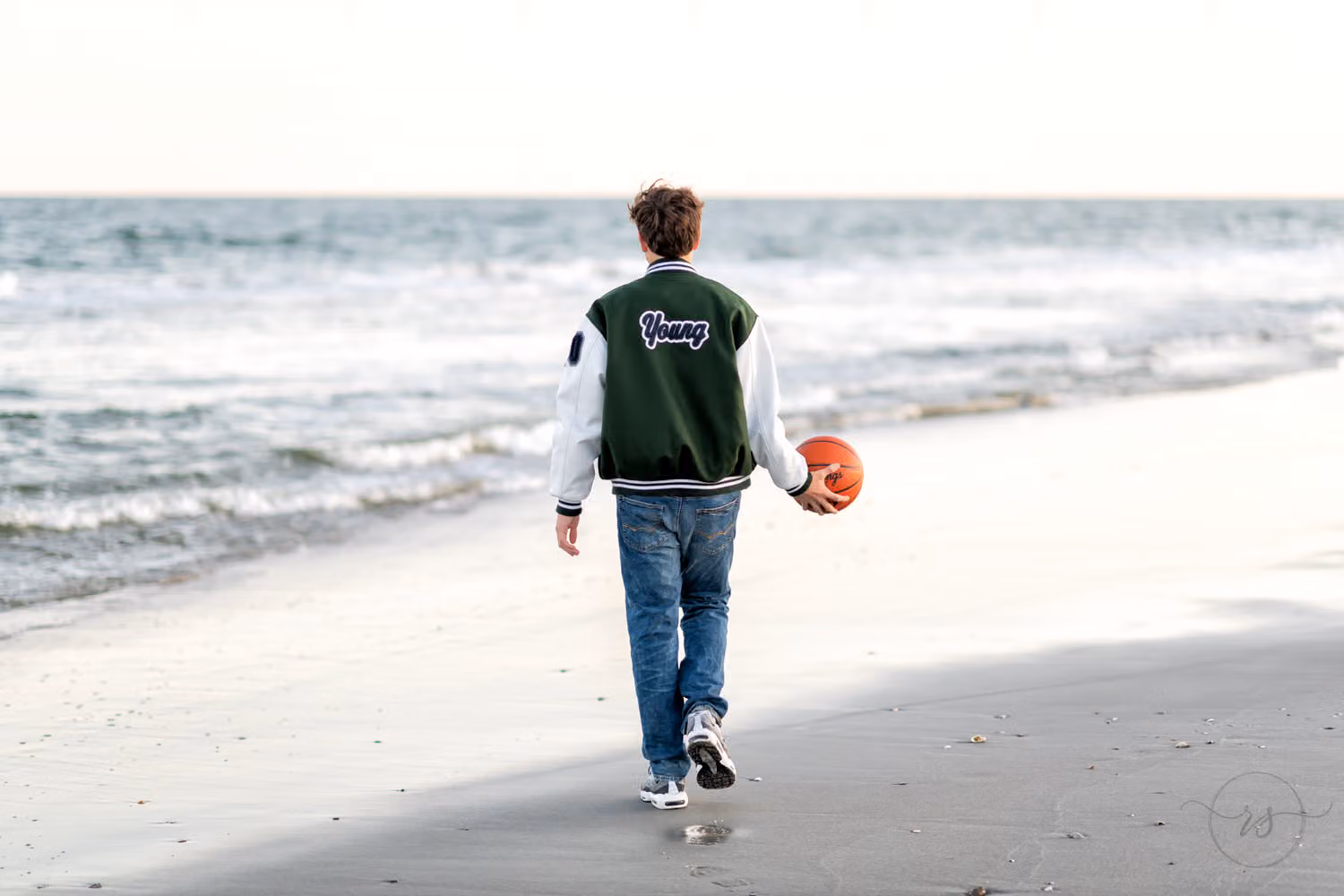 Senior in a letterman jacket walking away down the beach with a basketball at his side at Myrtle Beach State Park