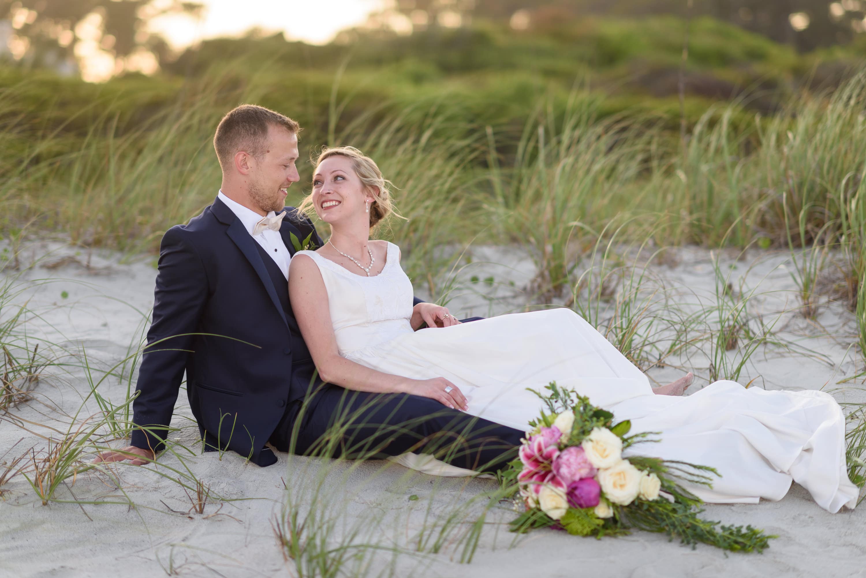 Bride smiling up at groom laying in dunes -  Grande Dunes Ocean Club