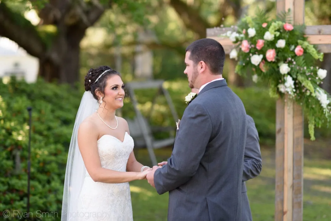 Bride and groom hold hands beside the floral cross during their vows at Upper Mill Plantation