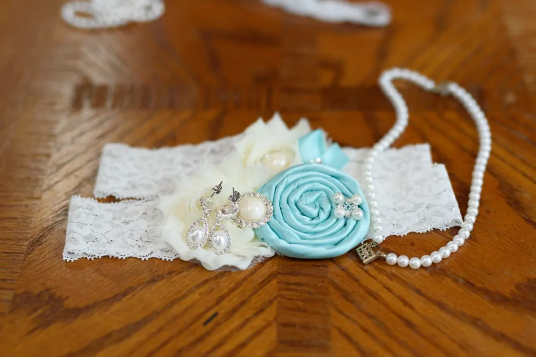 Lace garter with a teal rosette next to a pearl necklace on a wood table at Upper Mill Plantation