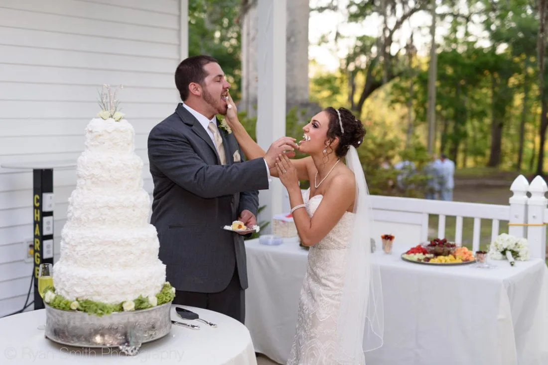 Bride and groom feed each other cake beside the tall white wedding cake at Upper Mill Plantation