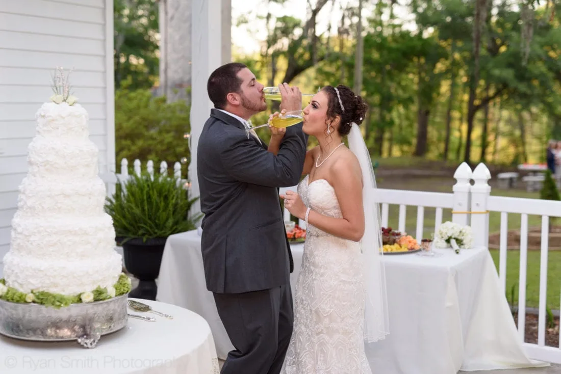 Groom takes a drink as the bride laughs beside the cake table at Upper Mill Plantation