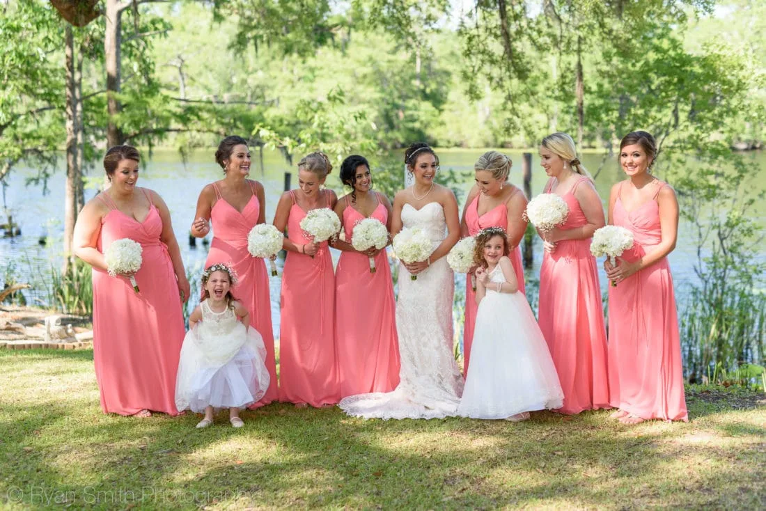 Bride with her bridesmaids in coral dresses and two flower girls by the lake at Upper Mill Plantation