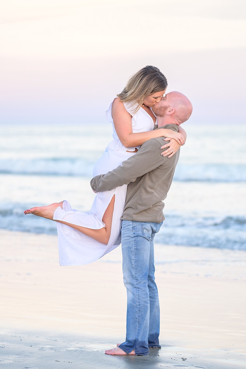 Lift into the air for a kiss in front of the ocean during engagement pictures