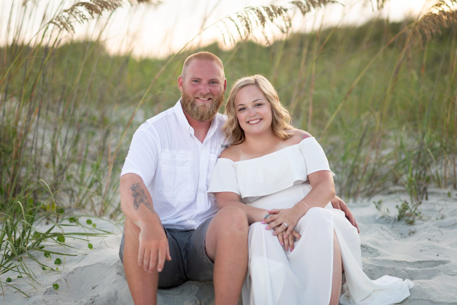 Couple in white sitting together in front of the sea oats at sunset -